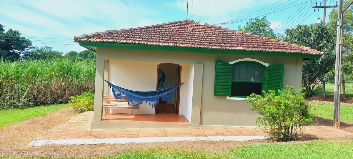 A charming house exterior is framed by green grass and light brown pathways. The building features a tiled roof and green shutters, with a blue hammock hanging on the porch. Nearby greenery and open fields create a peaceful rural setting.