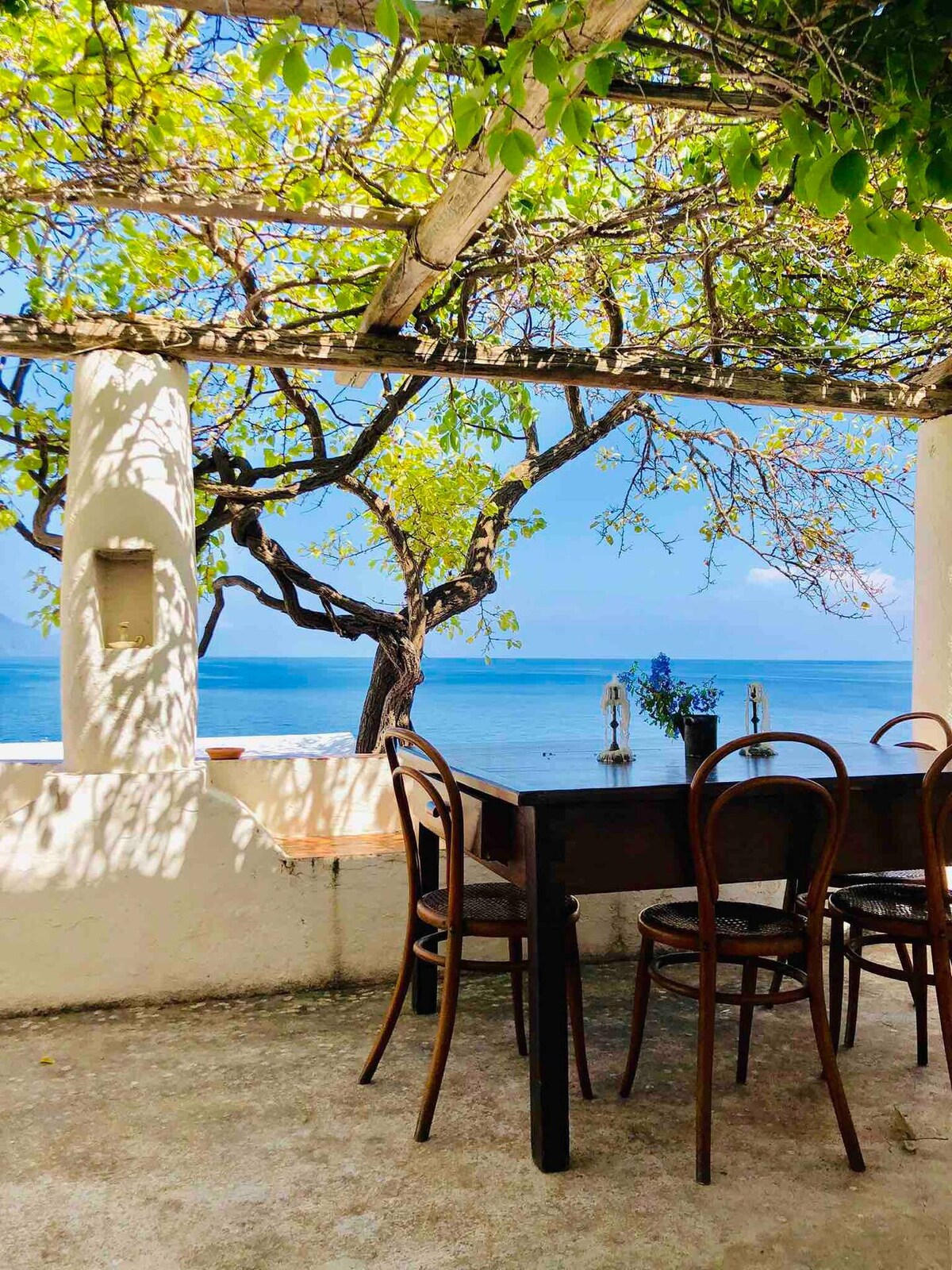 A dining area is set under a trellis adorned with lush greenery, providing shade while overlooking the sea. Wooden chairs surround a long table, and the tranquil water reflects the blue sky. Two lanterns are positioned decoratively on the table.