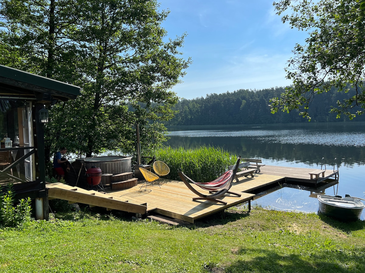 A wooden deck extends to the water's edge, featuring a hammock and colorful chairs for relaxation. A scenic view of the calm lake and lush greenery surrounds the area. A small boat is visible docked nearby, enhancing access to the water.