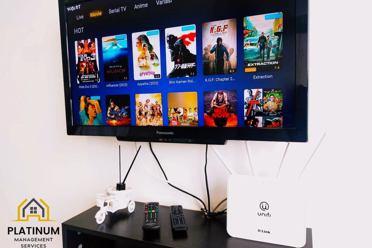 The image displays a wall-mounted television featuring a variety of movie options. A streaming device and remote controls are positioned below it. A white router stands nearby, along with a small decorative item, adding a touch of personality to the entertainment area.