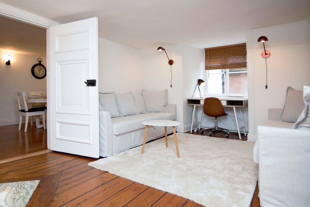 A bright living area features light-colored sofas, a small round table, and a stylish desk with a chair. Natural light filters through the window with wooden blinds, while warm wall sconces provide additional lighting. The space is accented by a soft, plush rug on the wooden floor.
