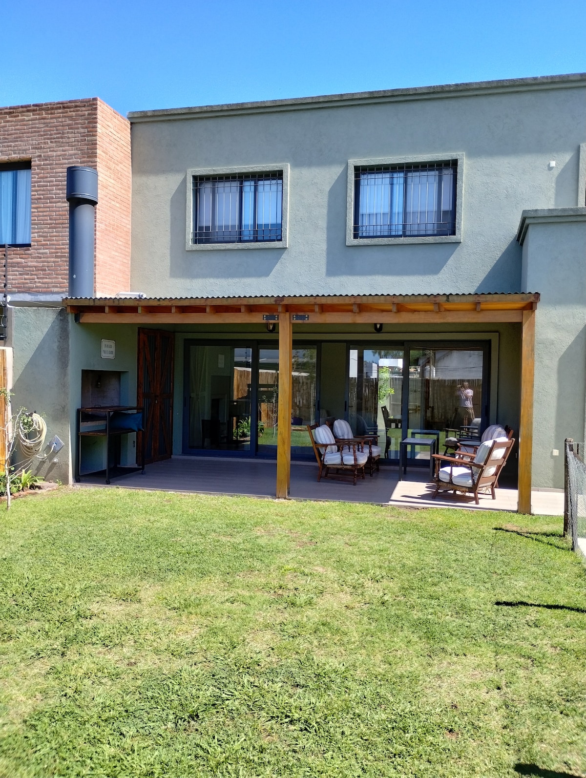 The outdoor patio area features wooden beams and a shaded porch with four comfortable chairs placed around a small table. Glass doors lead into the home, while a well-manicured lawn extends in front, providing a spacious outdoor environment.