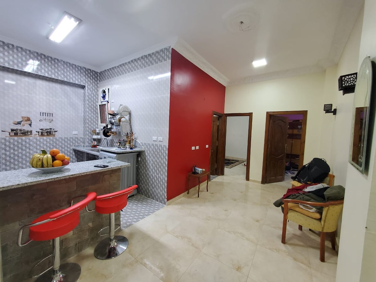 A clean kitchen area features modern bar stools in red, with a counter displaying fresh fruits. The wall behind is adorned with a patterned tile design, and a contrasting red wall adds a splash of color. A hallway leading to the living space is visible.