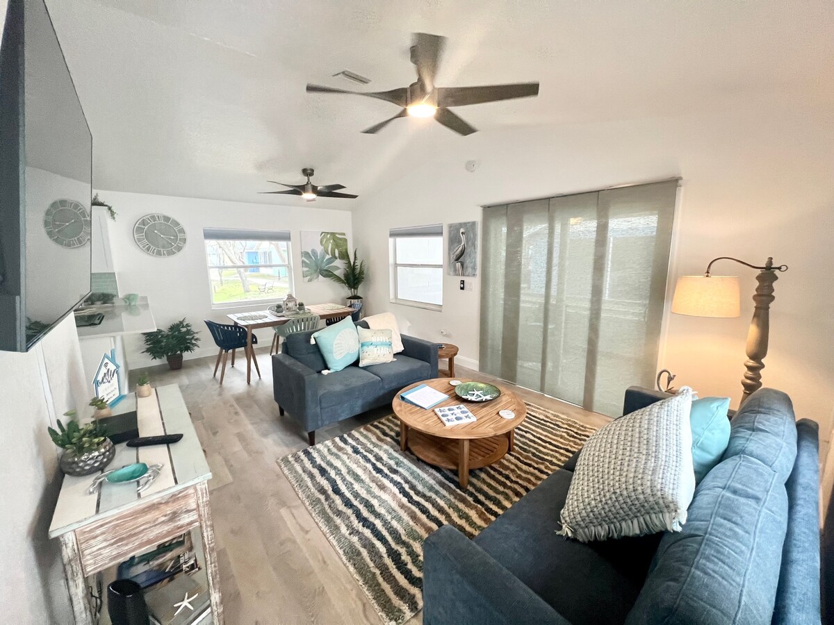 The living area features two comfortable sofas adorned with decorative pillows, positioned around a coffee table. A large flat-screen TV is mounted on the wall, while a cozy rug lays across the light wood flooring. Natural light filters through sliding glass doors, illuminating the space.