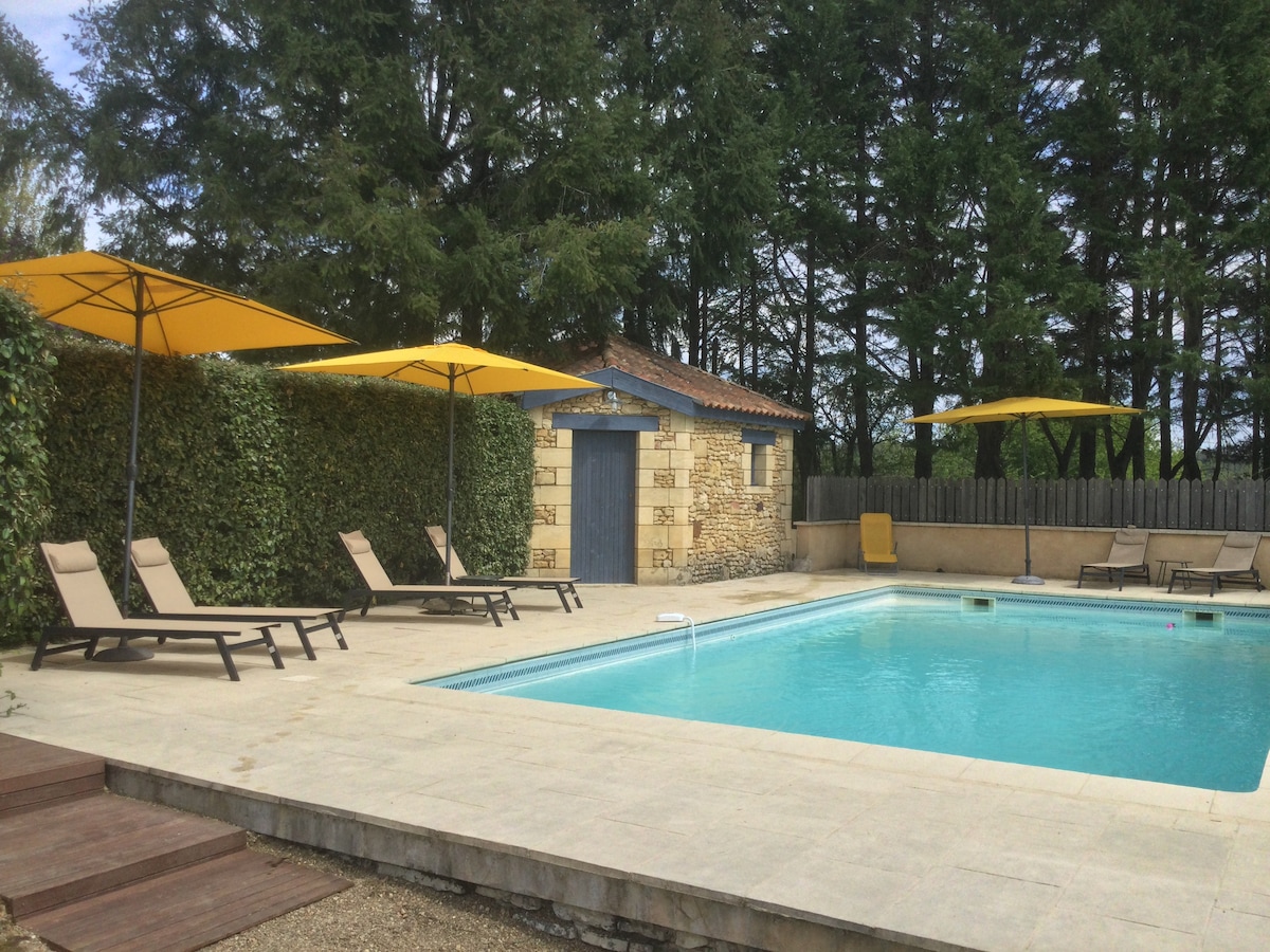 A spacious outdoor area features a swimming pool bordered by loungers under yellow umbrellas. Lush greenery provides privacy, while a quaint stone building with a blue door serves as a backdrop. Sunlight illuminates the tranquil scene, enhancing a relaxing environment.
