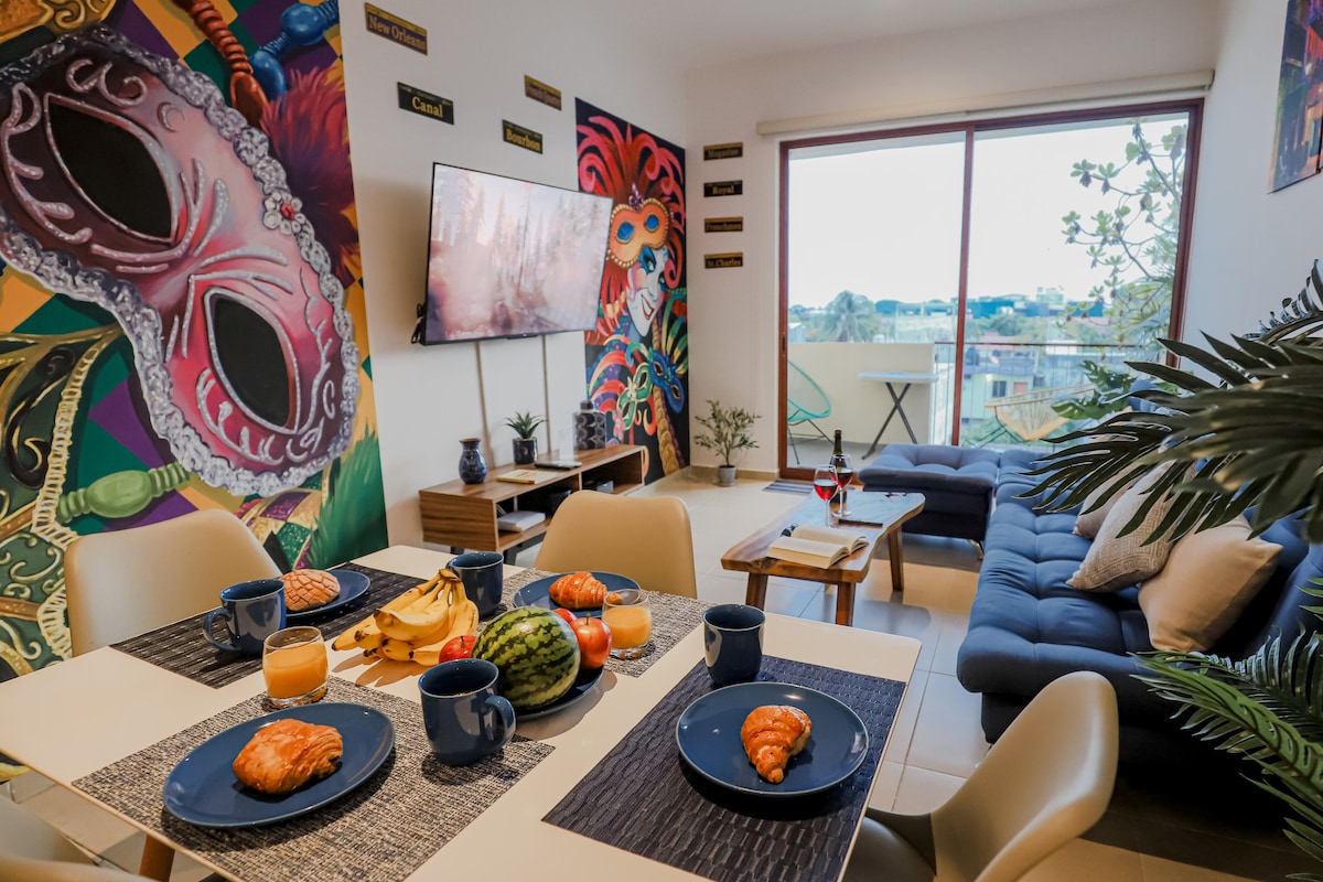 A dining area is set with plates of croissants and fresh fruit, accompanied by beverages. Colorful murals adorn the walls, enhancing the lively atmosphere. A large television is mounted in view of a cozy seating arrangement, with a balcony visible through expansive windows.