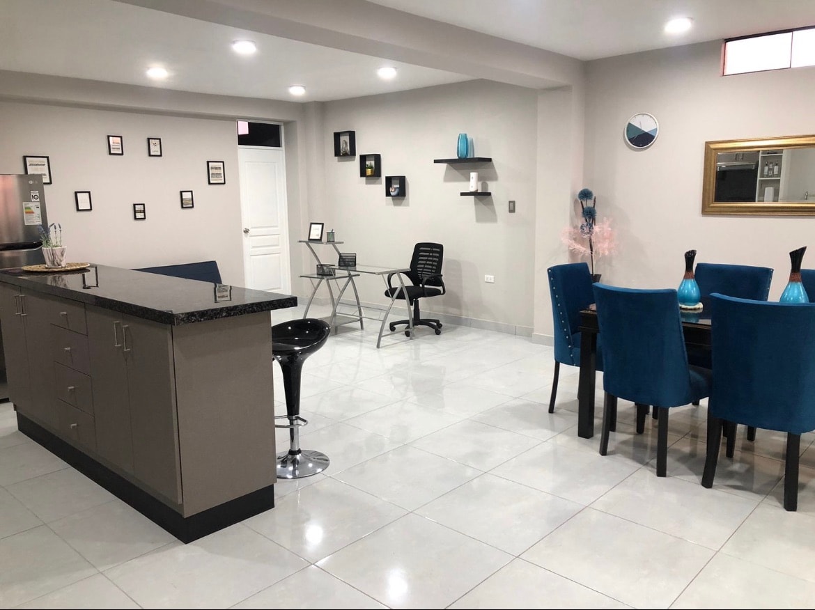 An open-plan living area is presented, featuring a contemporary kitchen island with additional seating. Blue upholstered chairs surround a dining table, while a workspace is positioned nearby with a desk and chair. Wall art and decorative elements are subtly displayed throughout the space.