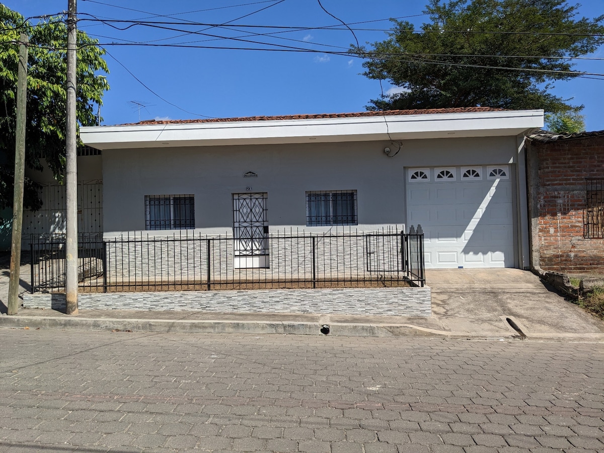 A single-story house is depicted with a light-colored exterior and a flat roof. A decorative wrought iron fence encloses the front yard, while a paved walkway leads to the entrance. The garage features a white door with multiple panels. Clear skies and greenery complement the setting.