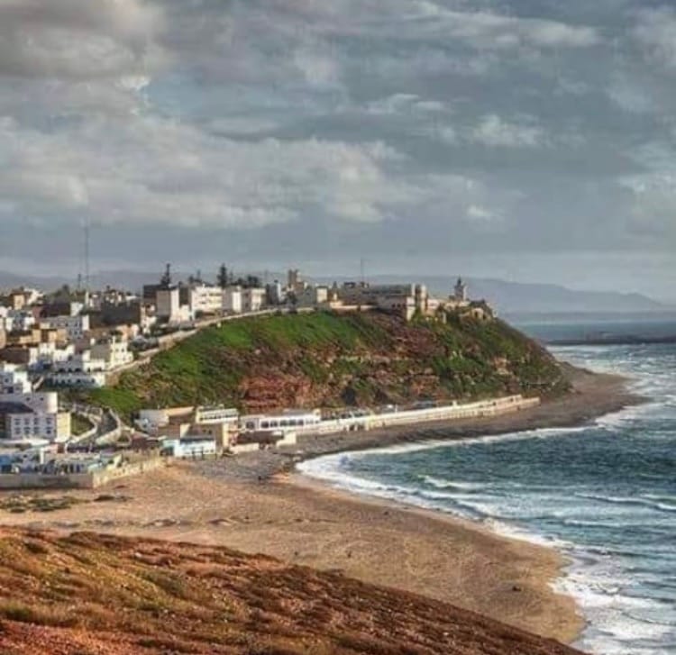 A scenic view of a coastal area is presented, featuring a curved sandy beach meeting the ocean. Low-lying buildings are visible along the coastline, situated on a hill that slopes gently towards the water. The sky is filled with soft clouds, enhancing the natural landscape.
