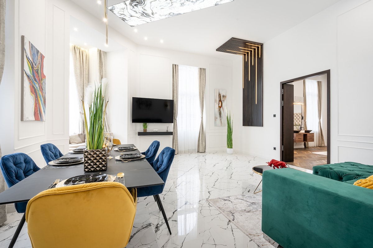 A bright living area features a dining table set for six and a spacious sofa in vibrant colors. Large windows with soft curtains allow natural light to fill the room, while contemporary artwork adds visual interest to the walls. A flat-screen TV is mounted for entertainment.
