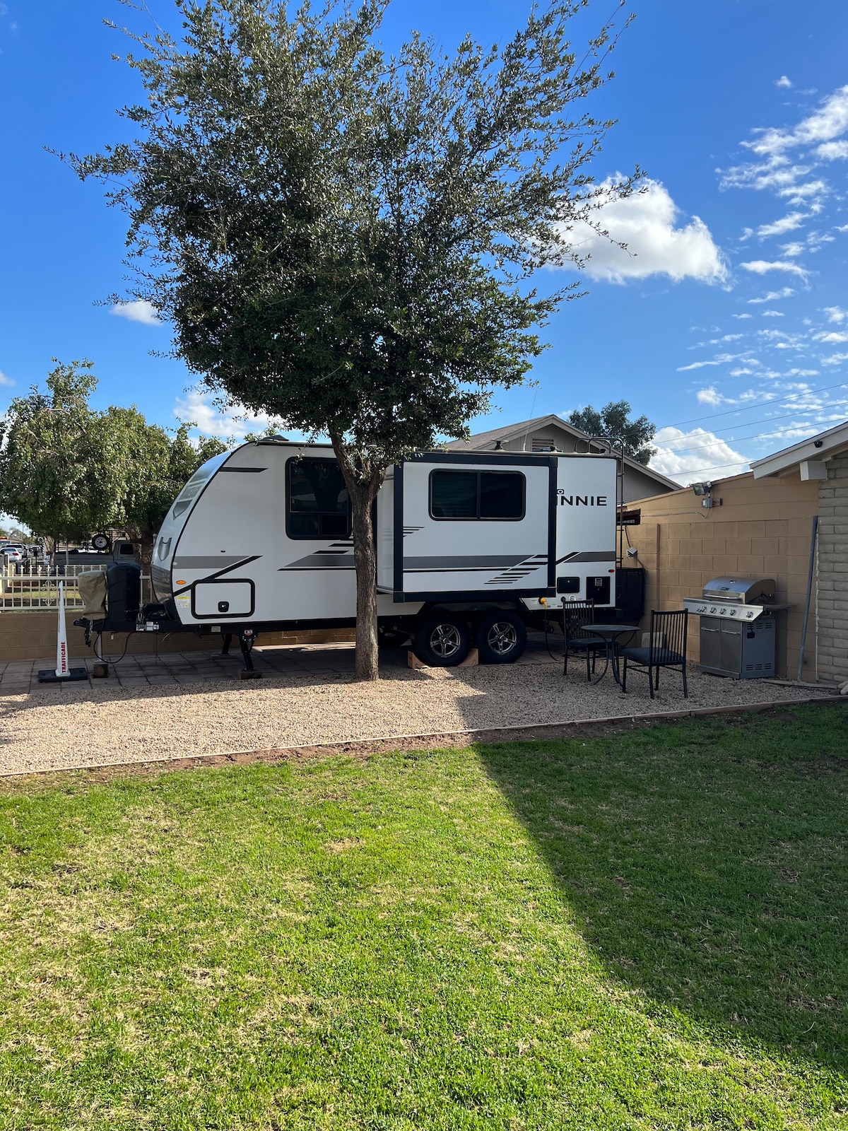 A camper rental is positioned in a shaded area, showcasing a sleek design with large windows. A small outdoor space includes a grassy area and gravel, accompanied by a patio table and chairs. A grill is visible nearby, providing an option for outdoor cooking.