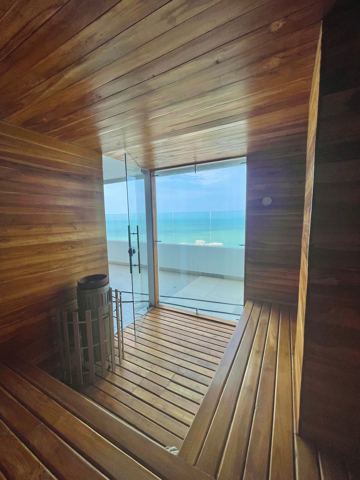 A serene sauna space features wooden paneling and a clear glass door that opens to a balcony. A small waste bin is positioned in the corner. Ocean views are visible through the glass, enhancing the calming environment.