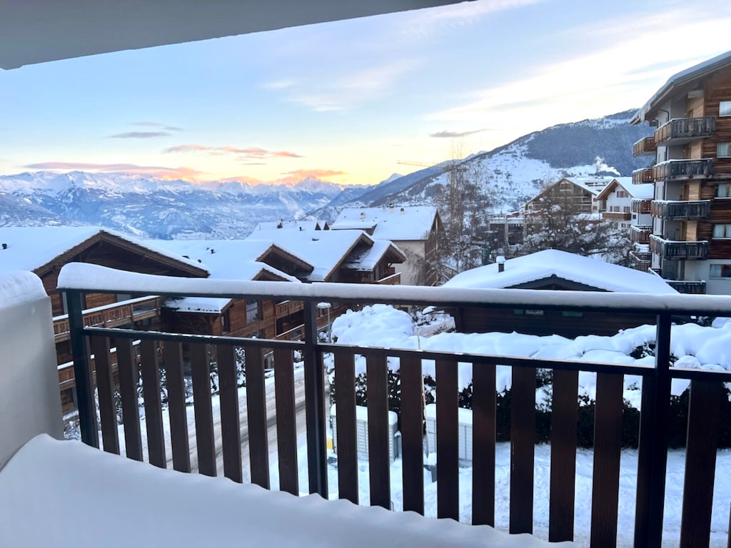 A balcony view captures snow-covered rooftops and the distant mountain range bathed in soft evening light. The landscape showcases a mixture of wooden chalets and snow, with the horizon revealing peaks under a pastel sky.