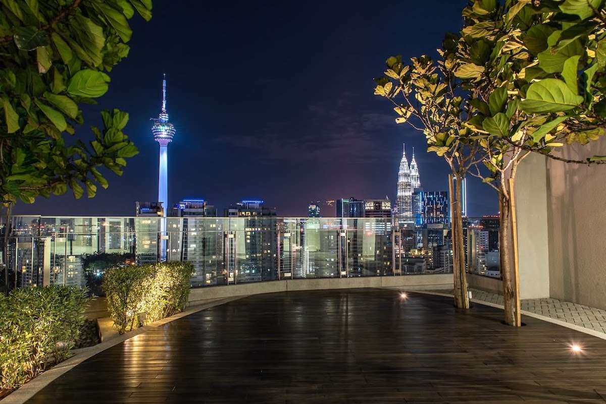 A spacious rooftop area is displayed, surrounded by greenery and offering views of the KL Tower and the Petronas Twin Towers illuminated at night. The wooden deck is complemented by city lights and a serene atmosphere, ideal for relaxation.