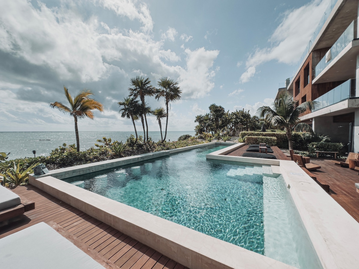 A serene pool area features a spacious infinity pool, surrounded by a wooden deck and tropical landscaping. The ocean can be seen in the background, framed by palm trees and lush greenery under a partly cloudy sky.