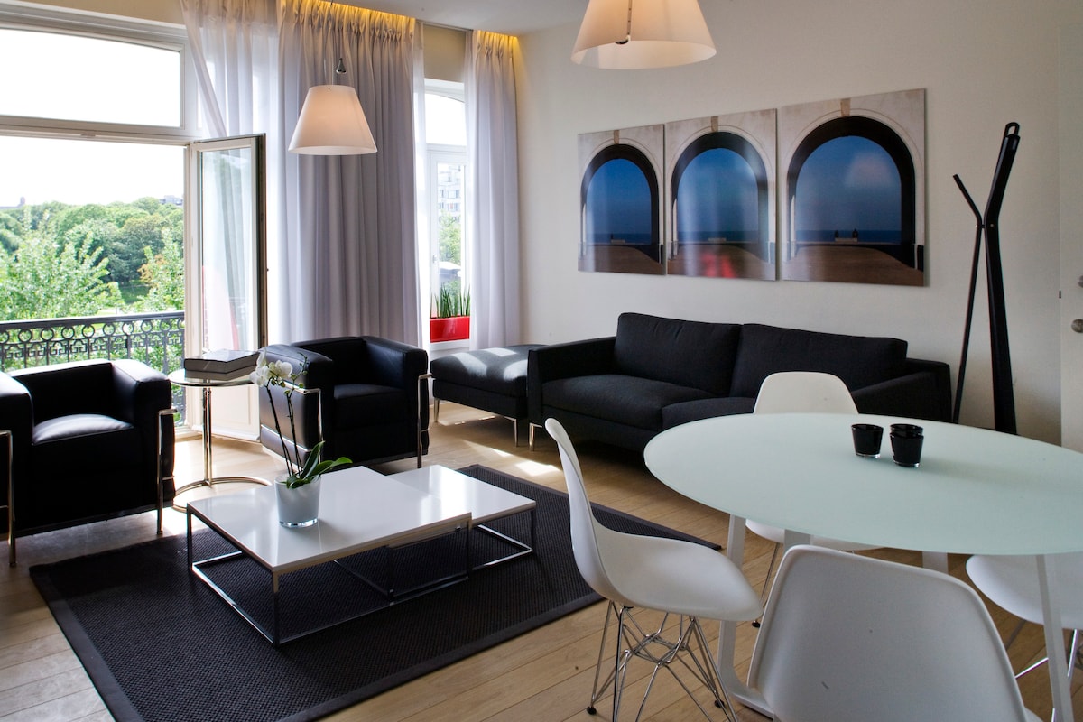 A bright living area is featured with large windows allowing natural light. Seating comprises a black sofa, two armchairs, and a dining table for four. Decorative art pieces adorn the wall, while a coffee table with a simple centerpiece adds to the welcoming ambiance.