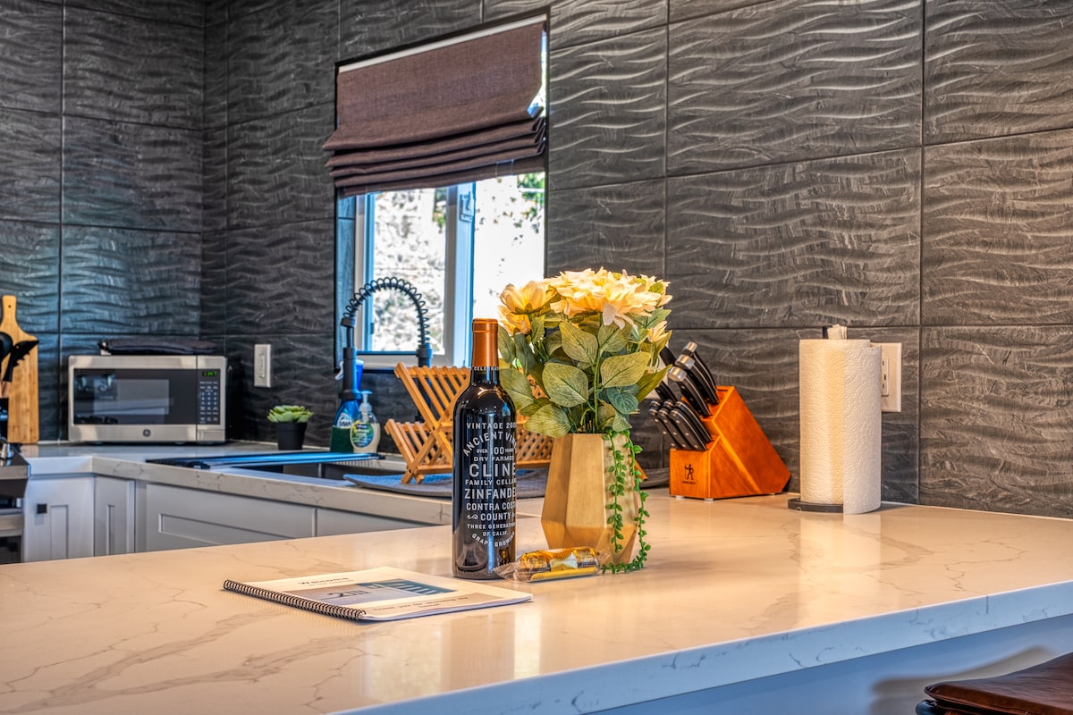 A modern kitchen features textured gray walls and a light quartz countertop. A bouquet of yellow roses is placed near a bottle of wine. Appliances, including a microwave and a knife set, are neatly arranged, contributing to a well-organized and functional space.