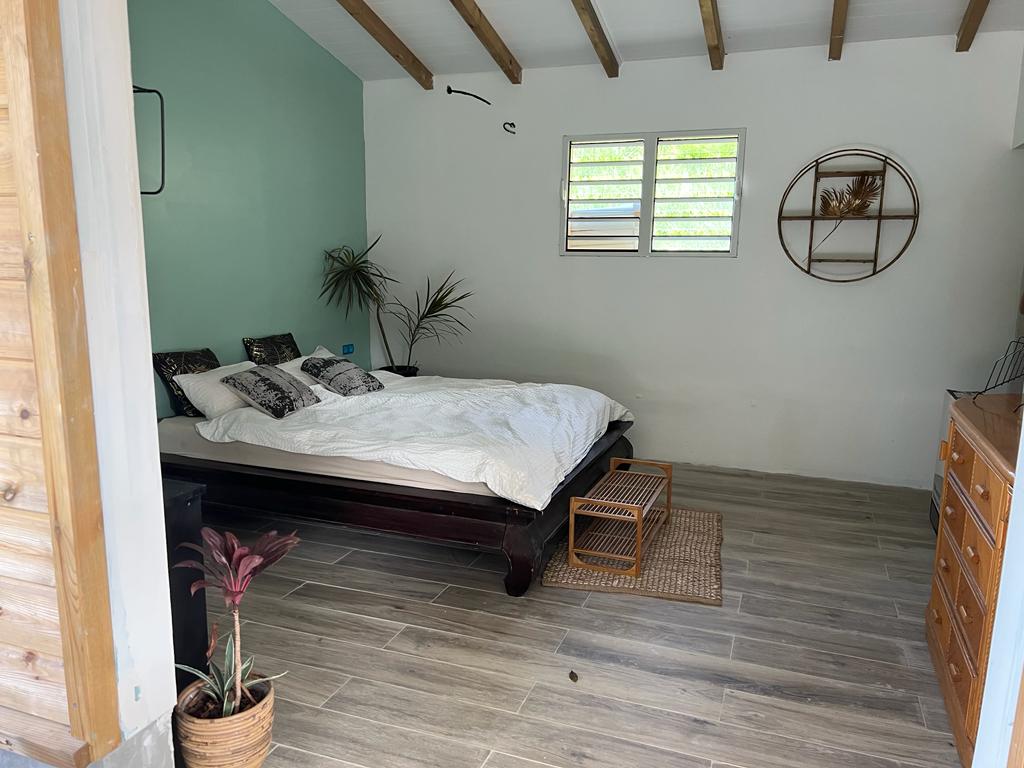 A tranquil bedroom is displayed, featuring a spacious bed with crisp white bedding. Natural light enters through partially open windows, highlighting light green walls. Minimalist decor is present, including a potted plant and a small wooden table, along with wooden beams adorning the ceiling.