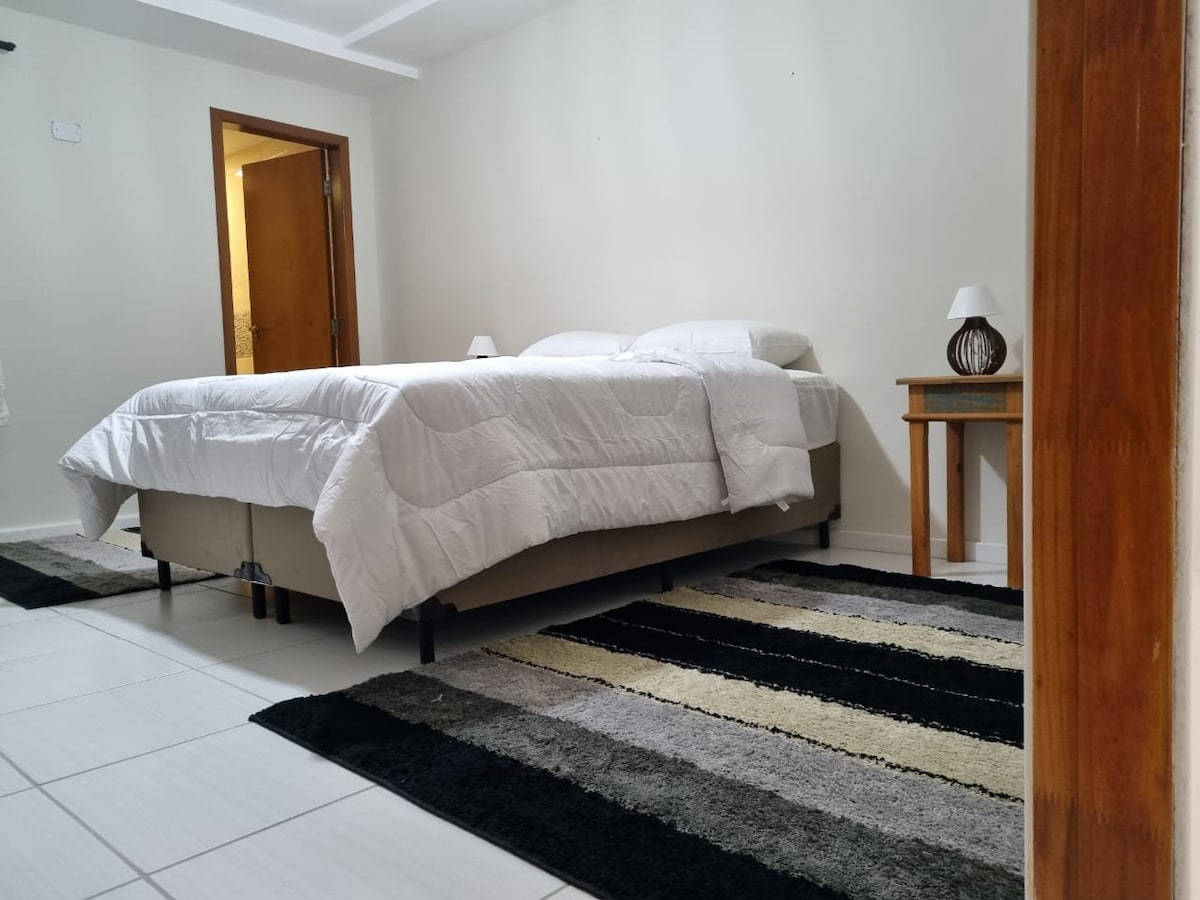 A spacious sleeping area features a large bed dressed in white bedding. A two-tone rug lies beneath, offering a soft contrast. A bedside table with a lamp is positioned alongside the bed, contributing to a bright and orderly environment.