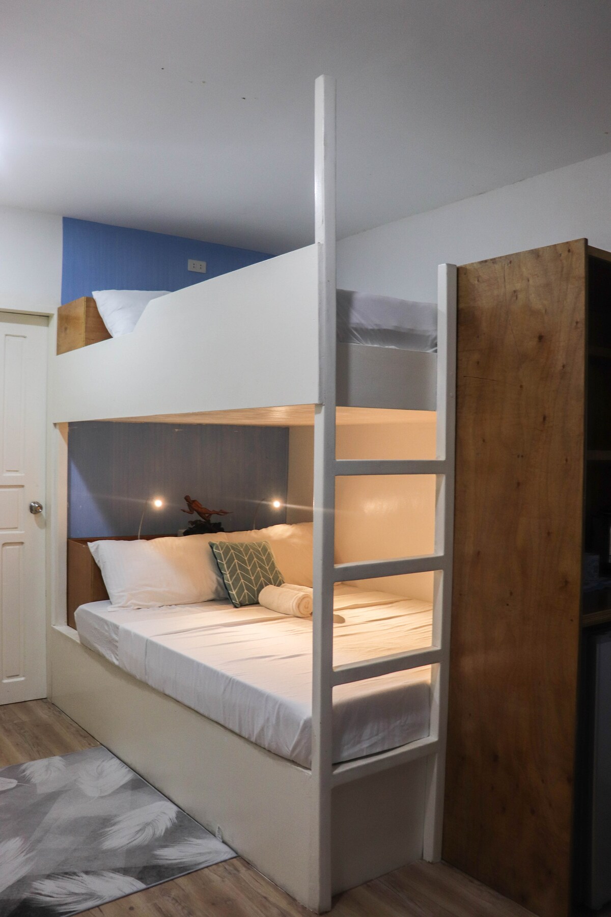 A private bedroom features a double bunk bed with a lower sleeping area showcasing crisp white linens and a decorative pillow. Soft lighting is mounted above each bed. A wooden partition provides storage space, enhancing the room's functionality while maintaining a clean and minimal aesthetic.