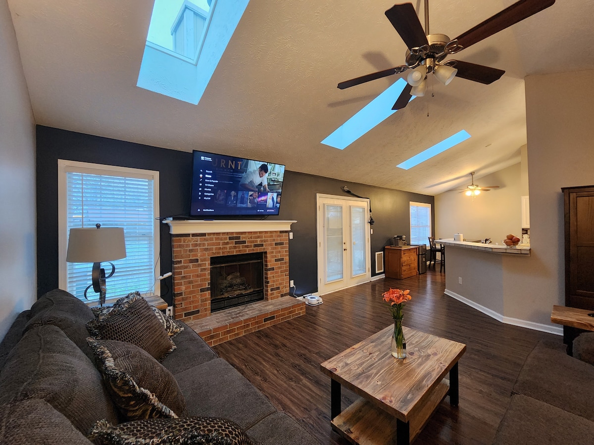 A spacious living area features a comfortable sectional sofa facing a large wall-mounted television. Natural light enters through two skylights and a set of glass doors leading to the outside. A coffee table complements the cozy setting, with a decorative vase of flowers placed atop.