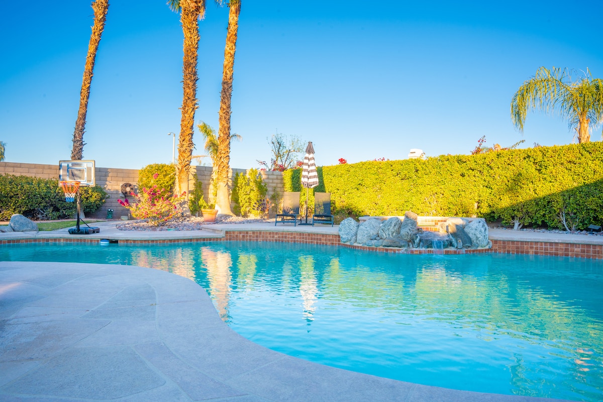 The outdoor space features a private swimming pool surrounded by lush landscaping. Palm trees and other greenery provide shade and privacy. A basketball hoop stands adjacent to the pool. A lounge chair can be seen beside a tranquil rock waterfall.