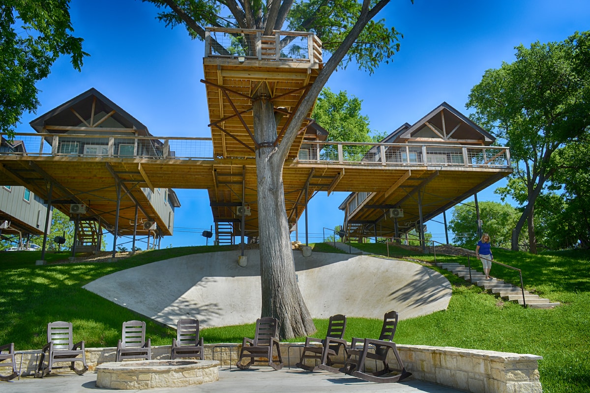 A multi-level cabin structure is set among lush greenery, elevated on wooden supports. Below the cabins, a patio area is seen, featuring multiple lounge chairs arranged in a semicircle. A large tree stands prominently, enhancing the natural landscape.