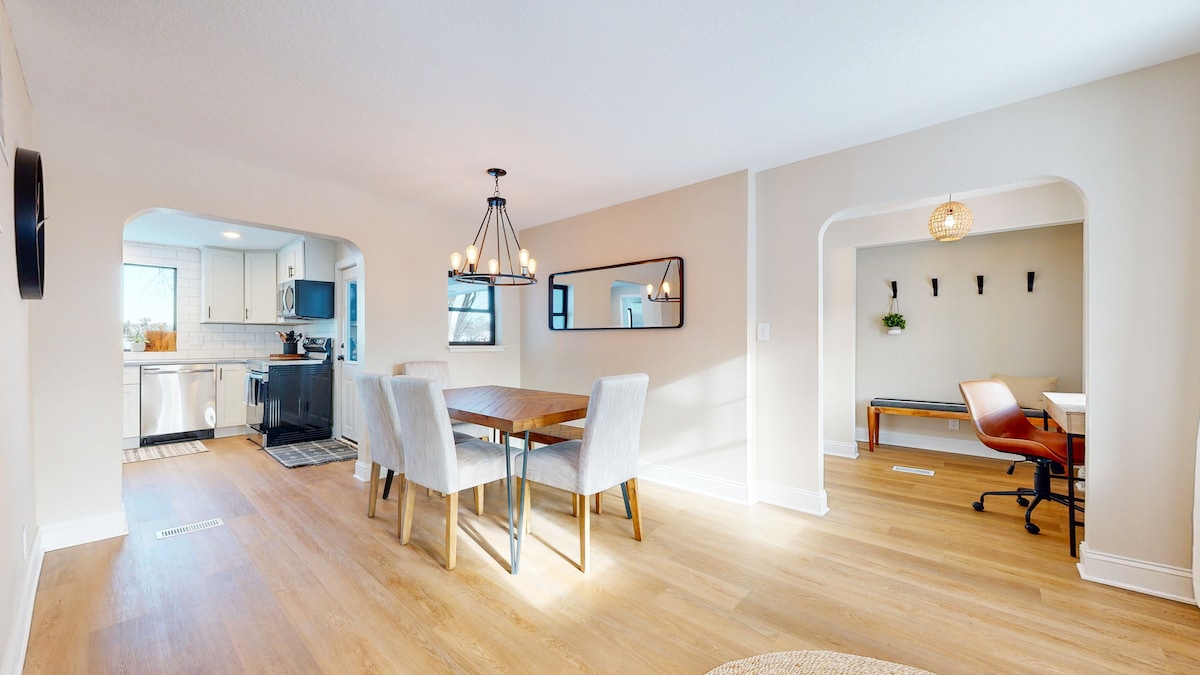 A bright dining area is highlighted by a wooden table surrounded by four fabric chairs. An adjoining space features a desk that provides a workspace. Natural light fills the room through windows, accentuating the light-colored walls and warm wooden flooring.