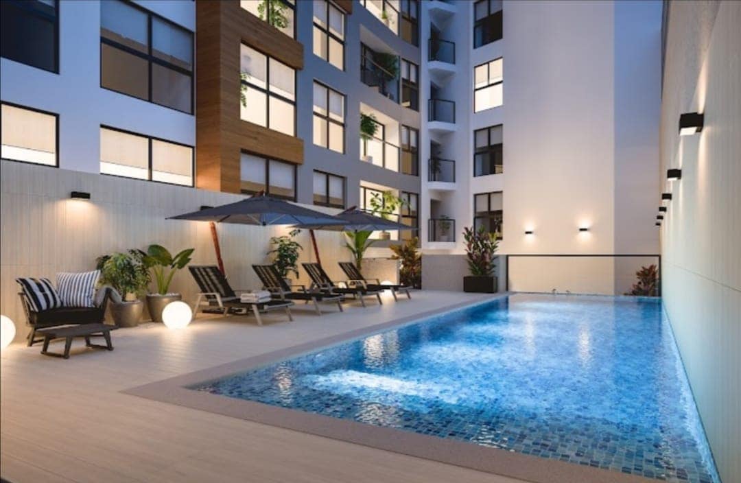 A sleek outdoor pool area features a mosaic-tiled swimming pool surrounded by comfortable lounge chairs. Umbrellas provide shaded seating, and lush greenery adds a refreshing touch. Natural light enhances the warm tones of the building's façade, creating a serene and inviting environment.