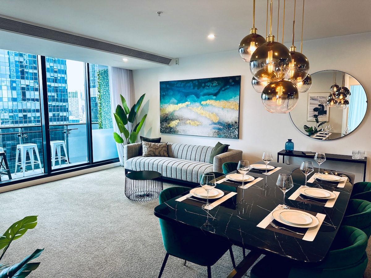 An open-plan living and dining area features a modern marble dining table with emerald velvet chairs. A stylish sofa, adorned with a patterned cushion, sits beneath a large artwork. Floor-to-ceiling windows allow for natural light and showcase the city views.