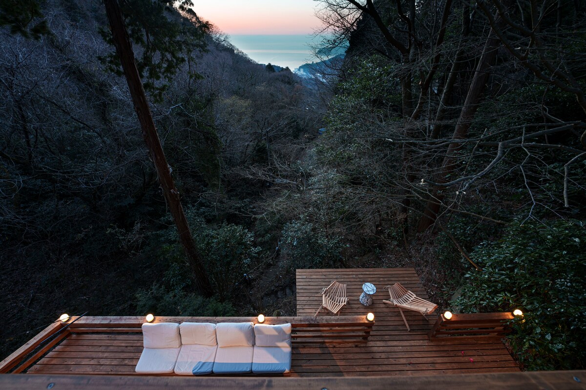 An outdoor deck area is visible, featuring a comfortable sofa and two lounge chairs. Soft lighting illuminates the space, surrounded by natural greenery and trees. The ocean can be seen in the distance, with the sky transitioning into twilight hues.
