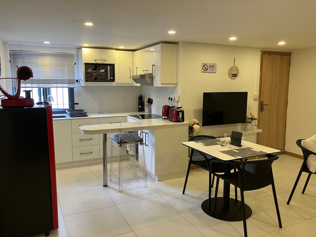 A modern kitchen area features sleek white cabinetry and stainless steel appliances. A transparent chair is positioned at a dining table set for two. A flat-screen television is mounted on the wall, accompanied by complementary kitchen accessories on the countertop.