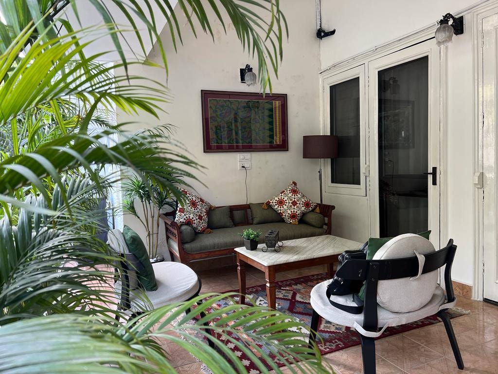 A cozy seating area is surrounded by lush green plants. A wooden coffee table sits in front of a comfortable couch adorned with patterned cushions. Glass doors offer a view into the room, while soft light is provided by a nearby lamp.