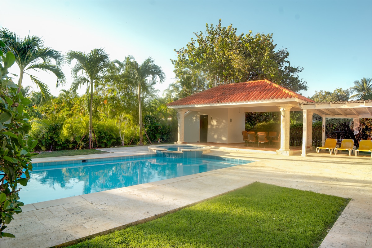 A spacious outdoor area features a sparkling blue pool surrounded by lush greenery. A shaded patio with seating is visible, along with a sunbathing area equipped with lounge chairs, creating a relaxing environment.
