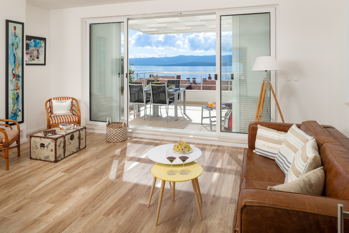 A bright living area features a brown leather sofa and a coffee table with fruit and drinks. Sliding glass doors lead to a spacious outdoor patio with dining furniture, offering views of the sea and sky. A decorative chair and art pieces add character.