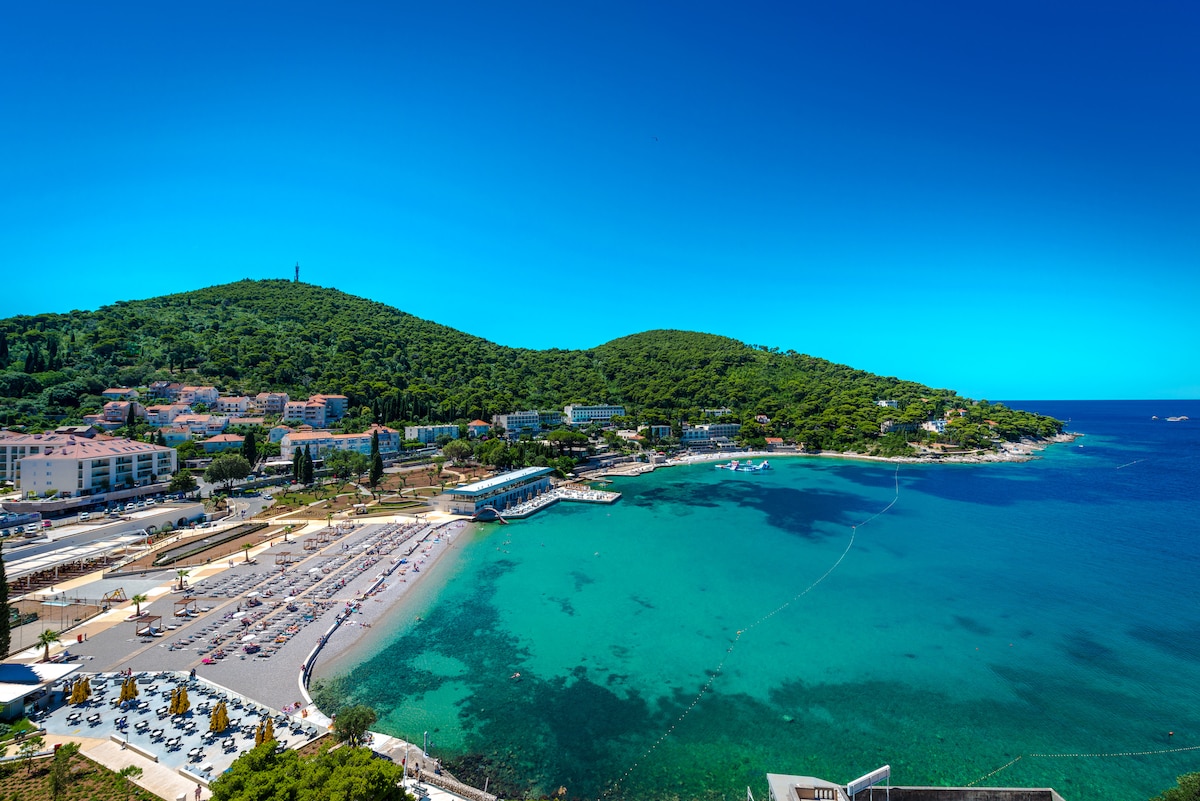 A panoramic view of a tranquil coastline is shown, featuring a sandy beach lined with sun loungers and umbrellas. Lush green hills rise in the background, under a clear blue sky. The serene turquoise water gently laps at the shore, creating a peaceful ambiance.