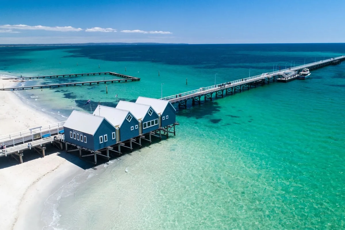 Aerial view of the beach showcasing a long wooden pier extending into clear turquoise waters. Three blue beach shacks sit alongside the shore, enhancing the coastal setting. Soft white sand lines the beach, with gentle waves lapping at the edge.