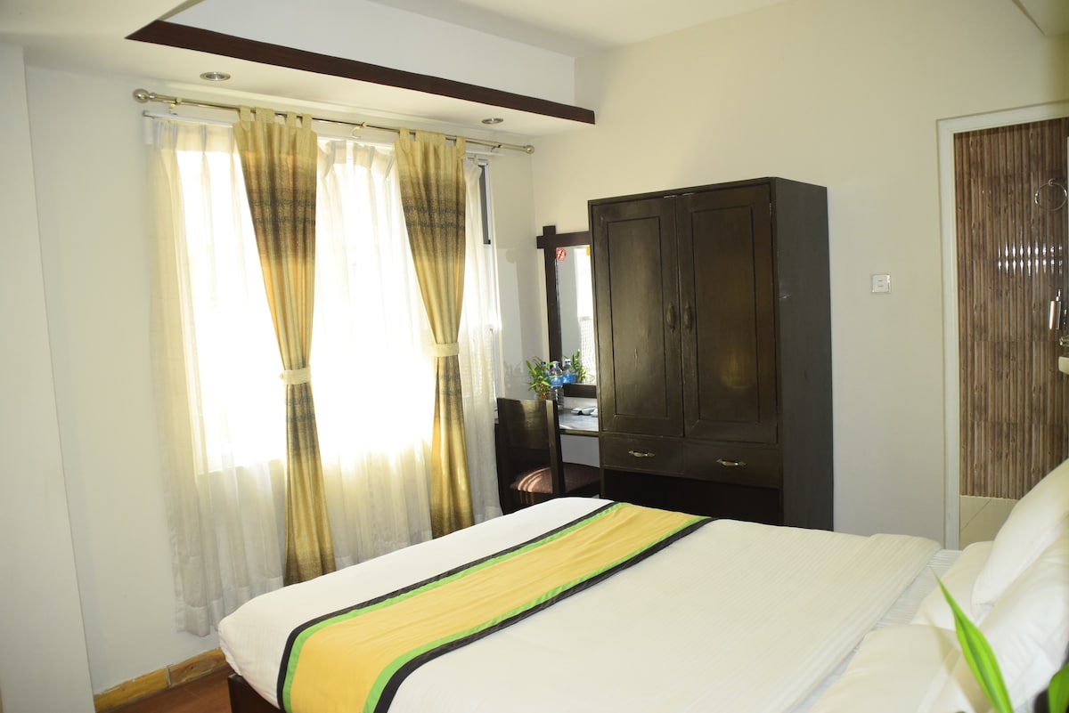 A well-lit room features a spacious bed adorned with a striped throw. Large curtains frame the window, allowing natural light to fill the space. A wooden cabinet and a reflective vanity are visible, enhancing the room's functionality and comfort.