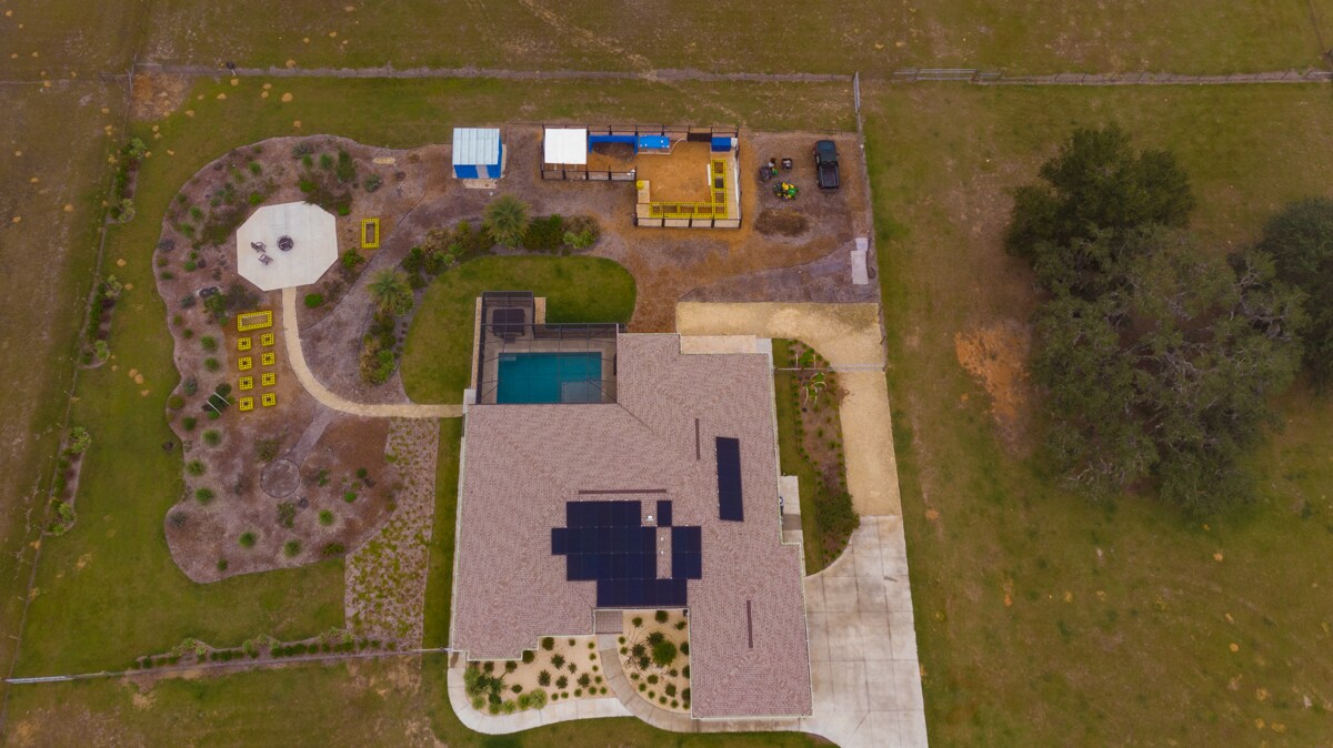 An aerial view showcases the property layout, featuring a spacious home with solar panels, a private pool, and an outdoor seating area. The landscaped garden includes a fire pit, surrounded by vibrant plant life, while large open fields extend beyond the fenced area.
