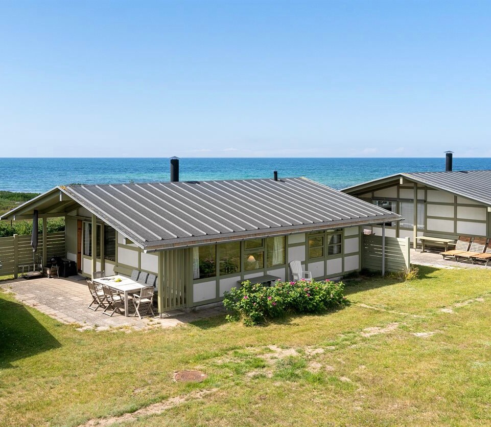 A serene view of a vacation house with a gray roof and white bindings, situated near the water. Multiple terraces are visible, surrounded by grassy areas that invite relaxation. The coastline can be seen in the background, enhancing the peaceful atmosphere of the location.