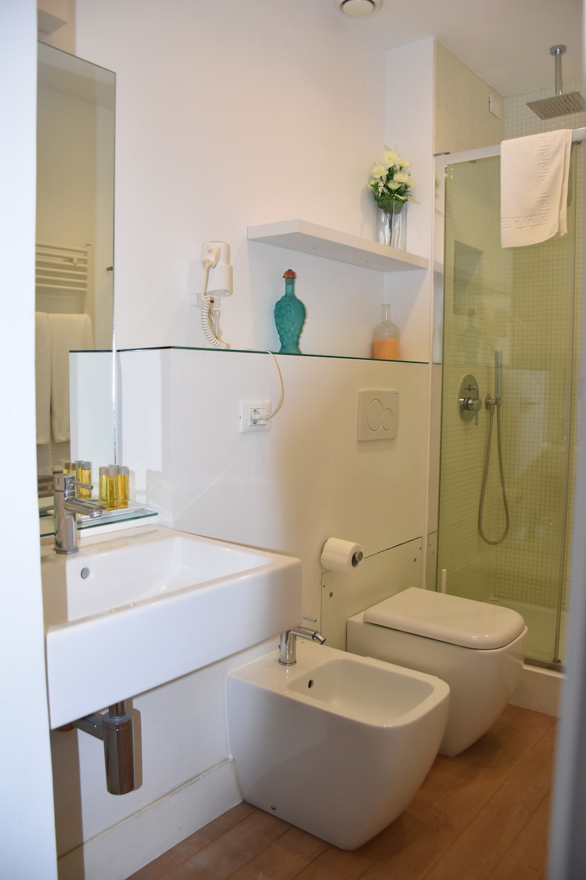A modern bathroom features a glass shower enclosure with a handheld showerhead and a wall-mounted shelf displaying a decorative vase. A white sink and bidet are positioned next to a compact toilet. Soft beige tones and natural light create a bright ambiance.