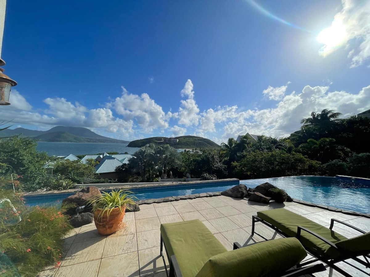 A serene pool area is framed by lush tropical greenery, showcasing a clear view of the Caribbean Sea and distant hills. Sunlight reflects off the water, while comfortable lounge chairs sit invitingly alongside the pool, creating a peaceful retreat.