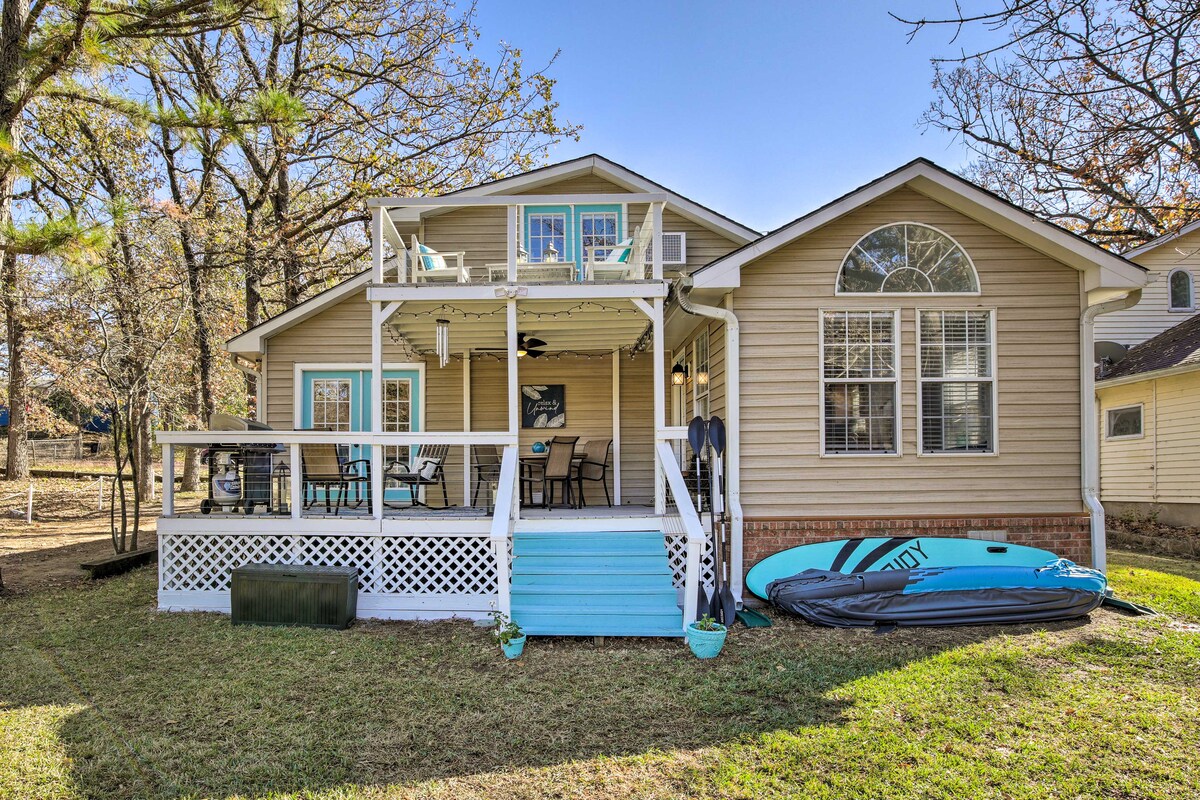 The exterior of the house showcases a welcoming front porch with a blue floor. Multiple outdoor seating options are visible, along with a kayak positioned against the wall. The well-maintained lawn surrounds the home, framed by trees and natural greenery.