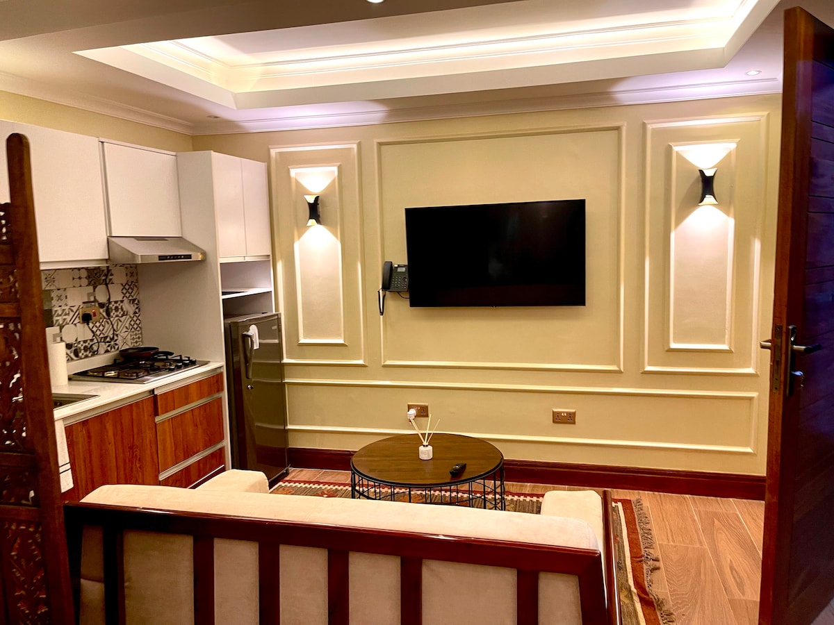 A modern studio is shown, featuring a comfortable seating area with a light-colored sofa next to a circular coffee table. A large wall-mounted television is visible, complemented by stylish wall sconces illuminating the room. The kitchenette, equipped with stainless steel appliances, is seen in the background.