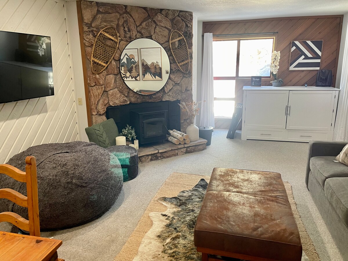 A cozy living room is highlighted by a stone fireplace and a large round mirror that reflects natural light. A super comfy LoveSac bean bag chair and a leather ottoman provide ample seating. A wooden dining table is showcased alongside a modern sideboard against the wall.