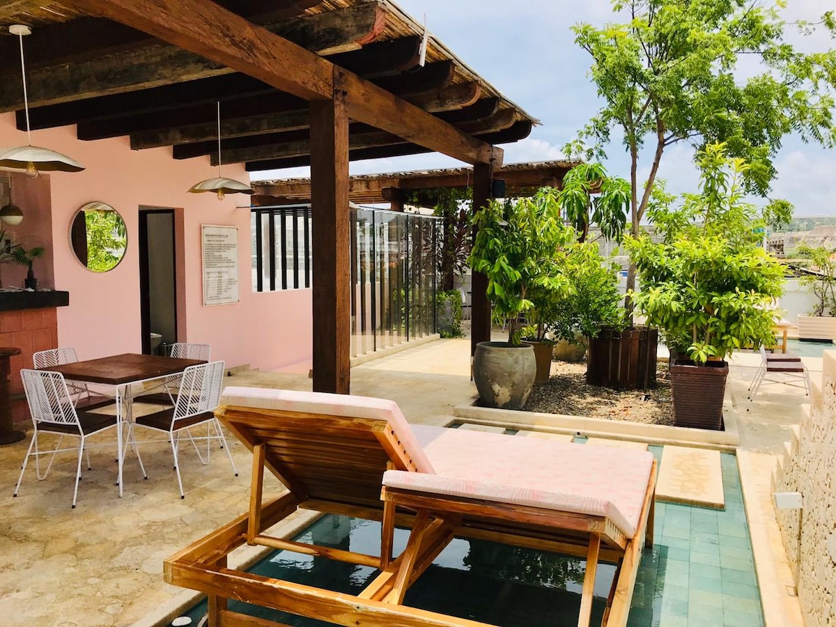 A spacious outdoor area is featured, showcasing a wooden lounge chair positioned by a small pool. Surrounded by greenery, the space includes a dining table with four chairs and a rustic wooden ceiling, creating an inviting environment for relaxation and dining.