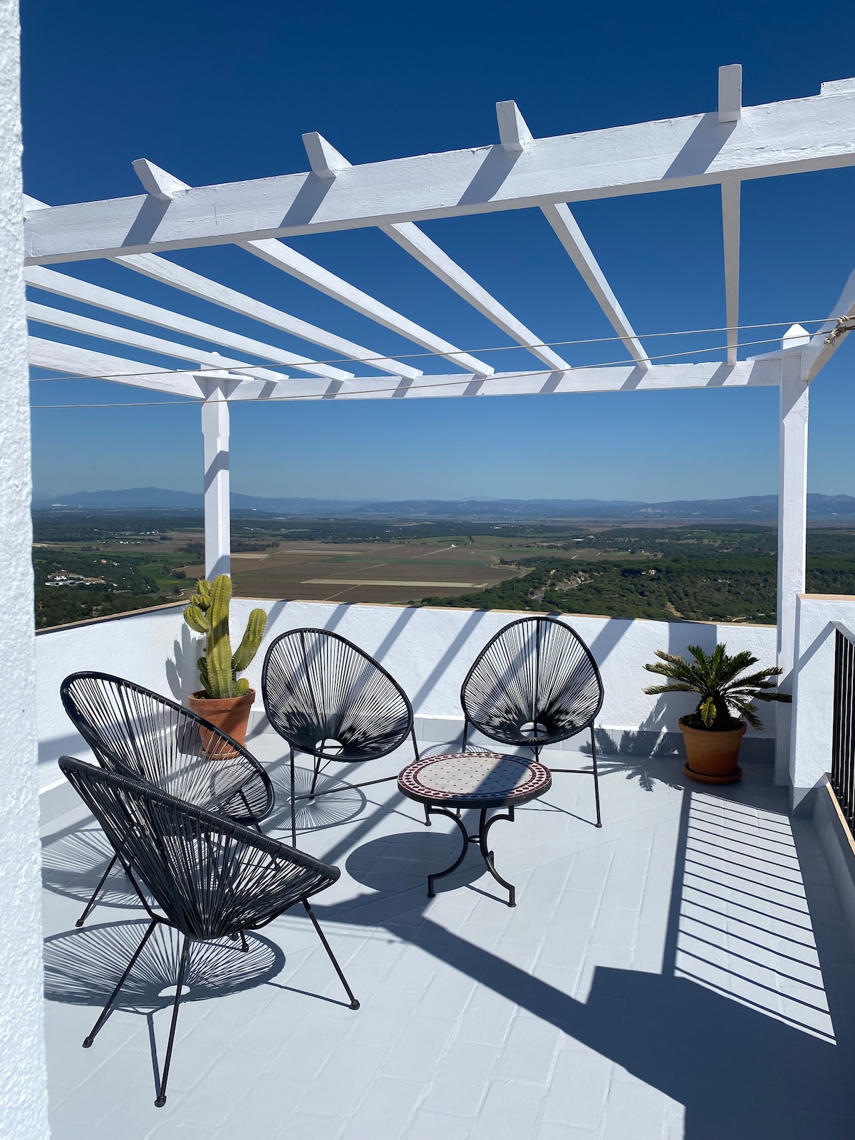 An open terrace features a white pergola casting shadows on the floor. Four black wire chairs surround a small circular table, while potted plants add greenery. The expansive view showcases fields and distant mountains under a clear blue sky.