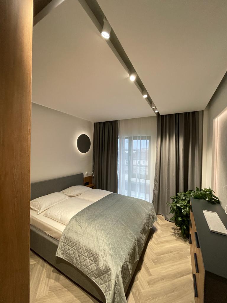 A modern bedroom is furnished with a spacious bed covered in neutral linens. Soft lighting highlights the minimalistic design. Large windows covered by sheer curtains allow natural light to fill the room, while a touch of greenery is seen in the corner.