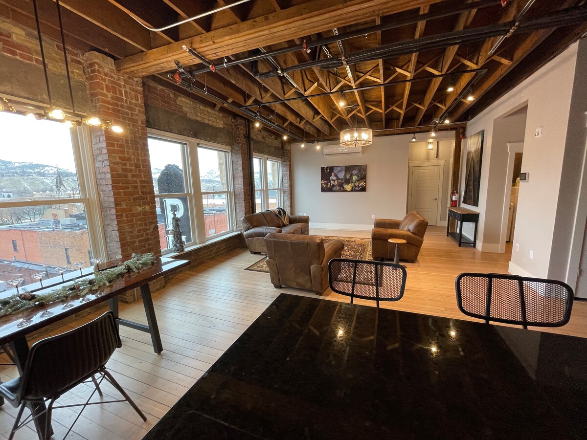 An open living area features exposed brick walls and wooden beams, enhancing the historic charm. A spacious seating arrangement includes a leather sofa and armchairs. Large windows provide ample natural light and views of the surroundings. A dining table is set nearby on polished wood flooring.