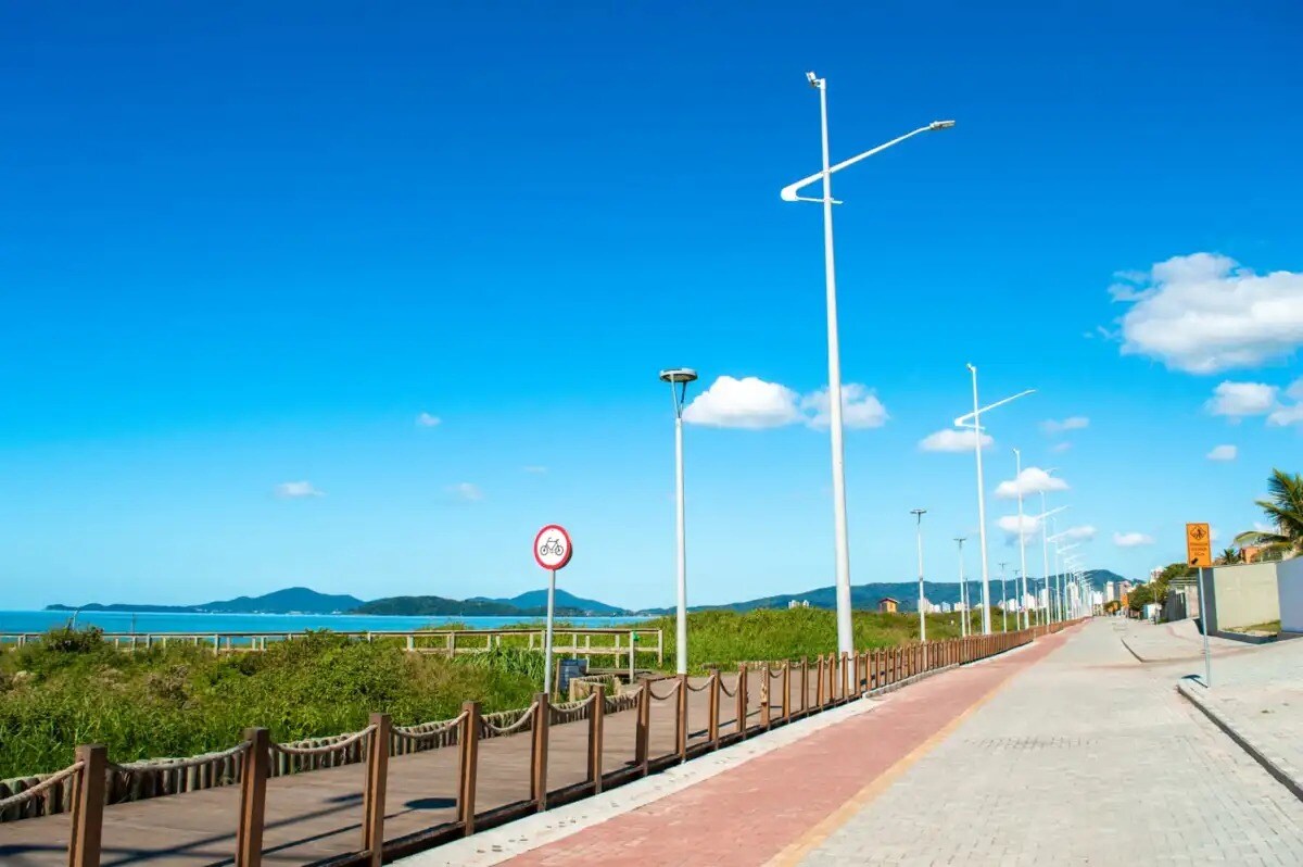 A wide coastal path is visible, lined with wooden railings. On one side, greenery leads to the shoreline, while the ocean is seen in the background under a clear blue sky. Streetlights dot the pathway, creating a well-lit environment for pedestrians.